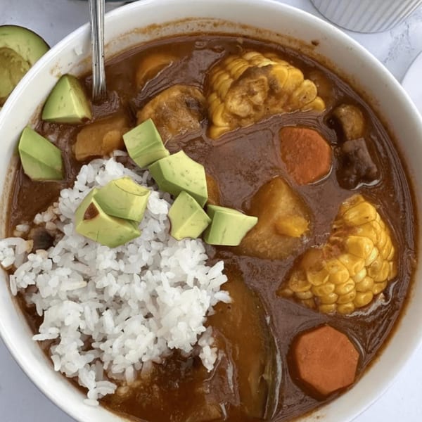 Hearty Puerto Rican stew with corn, carrots, avocado, and white rice in a white bowl.