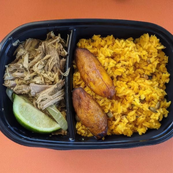 Puerto Rican meal with shredded pork, yellow rice, fried plantains, and a lime wedge in a black container.
