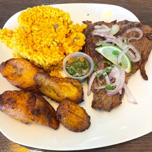 Puerto Rican steak with yellow rice, fried plantains, and a side of green sauce.