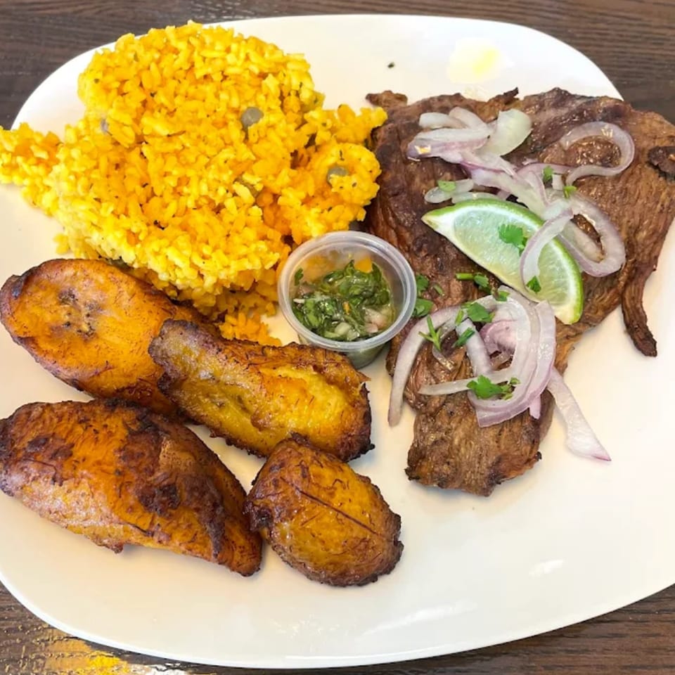 Puerto Rican steak with yellow rice, fried plantains, and a side of green sauce.